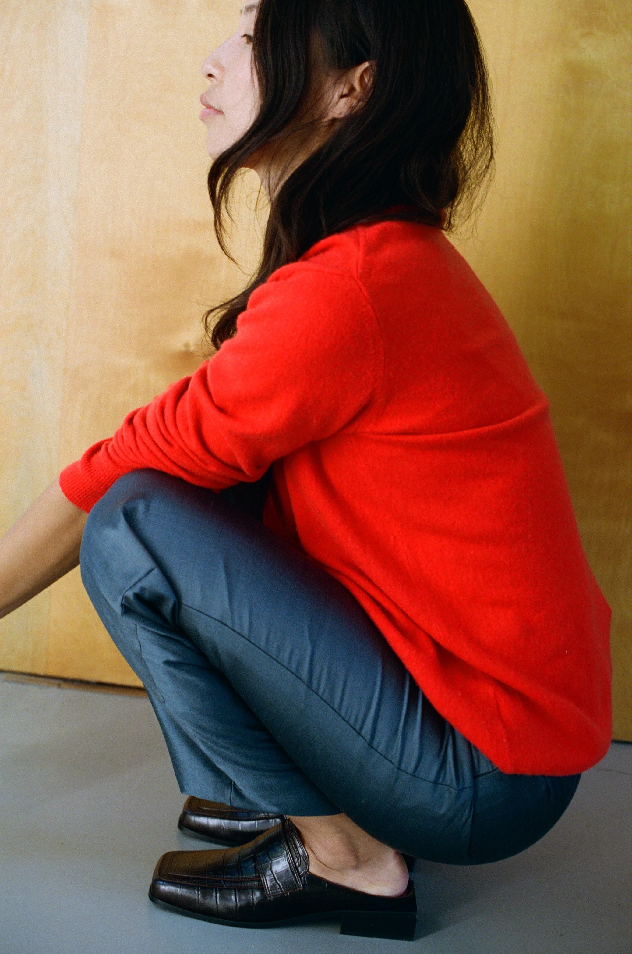 A person squatting sideways against a wooden wall, wearing a bright red sweater, dark trousers, and dark brown square toe slip-on loafers from SCOTTI. Their long, wavy hair drapes over their shoulder as they look thoughtfully into the distance.