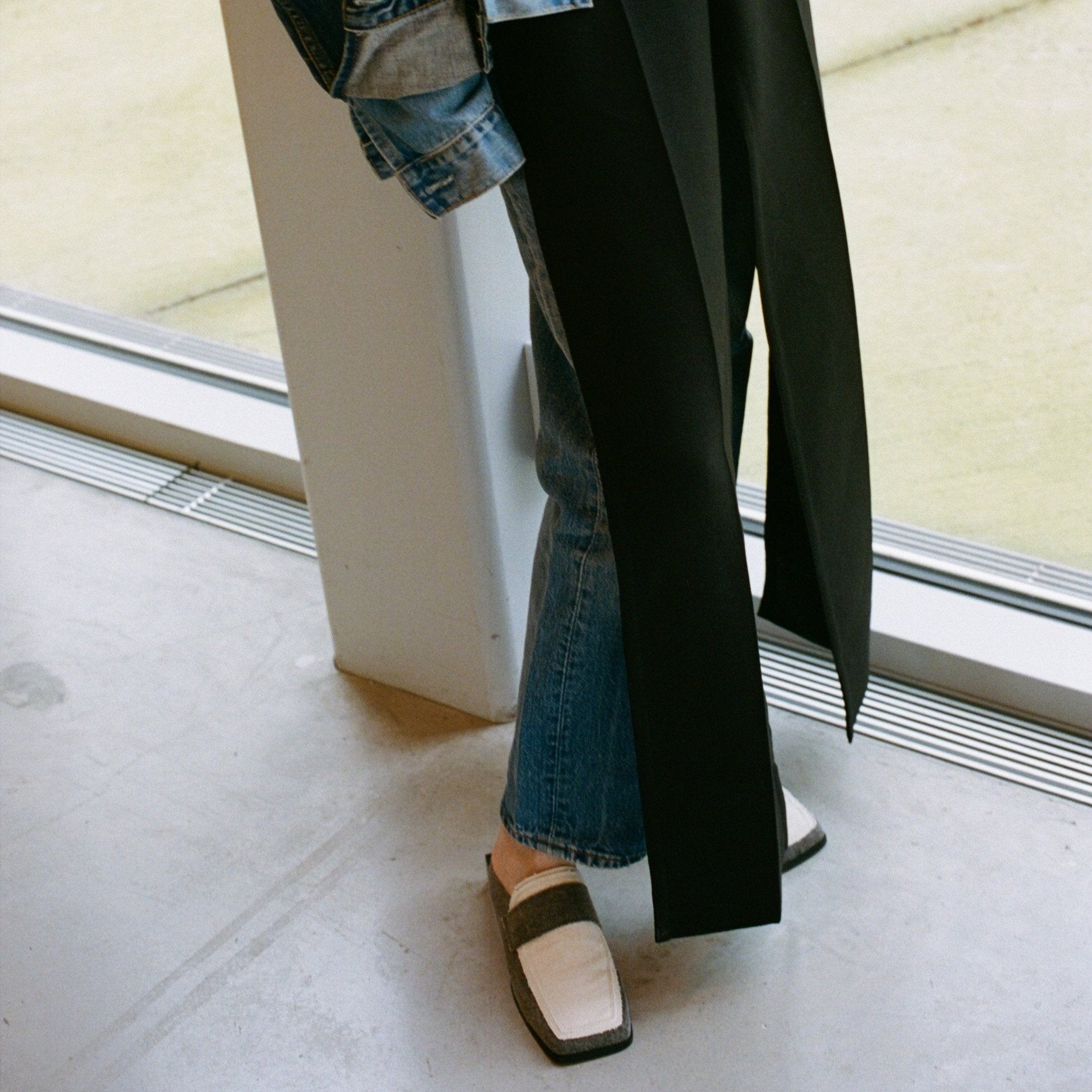 Lower half of a person wearing layered black pants over jeans with a rolled-up denim jacket, styled with SCOTTI square toe slip-on loafers. A bold designer footwear look, perfect for trend-forward summer outfits, captured by a window on a concrete floor.