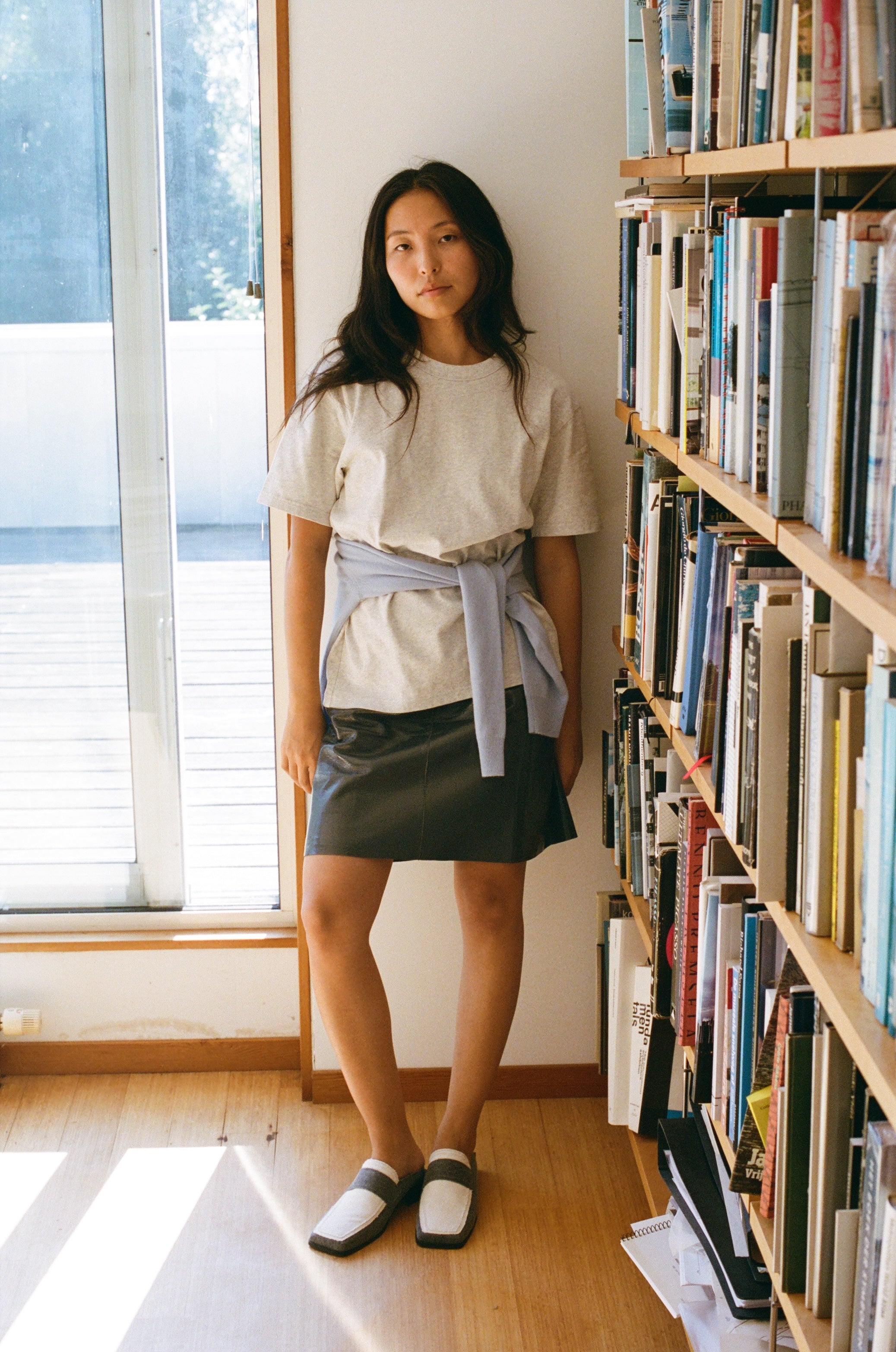 A person with long dark hair stands by a bookshelf in a sunlit room. She is wearing a light grey shirt with a sweater tied around the waist, a grey skirt, and white and grey denim vegan SCOTTI loafer with a square toe. A window on the left lets in natural light.