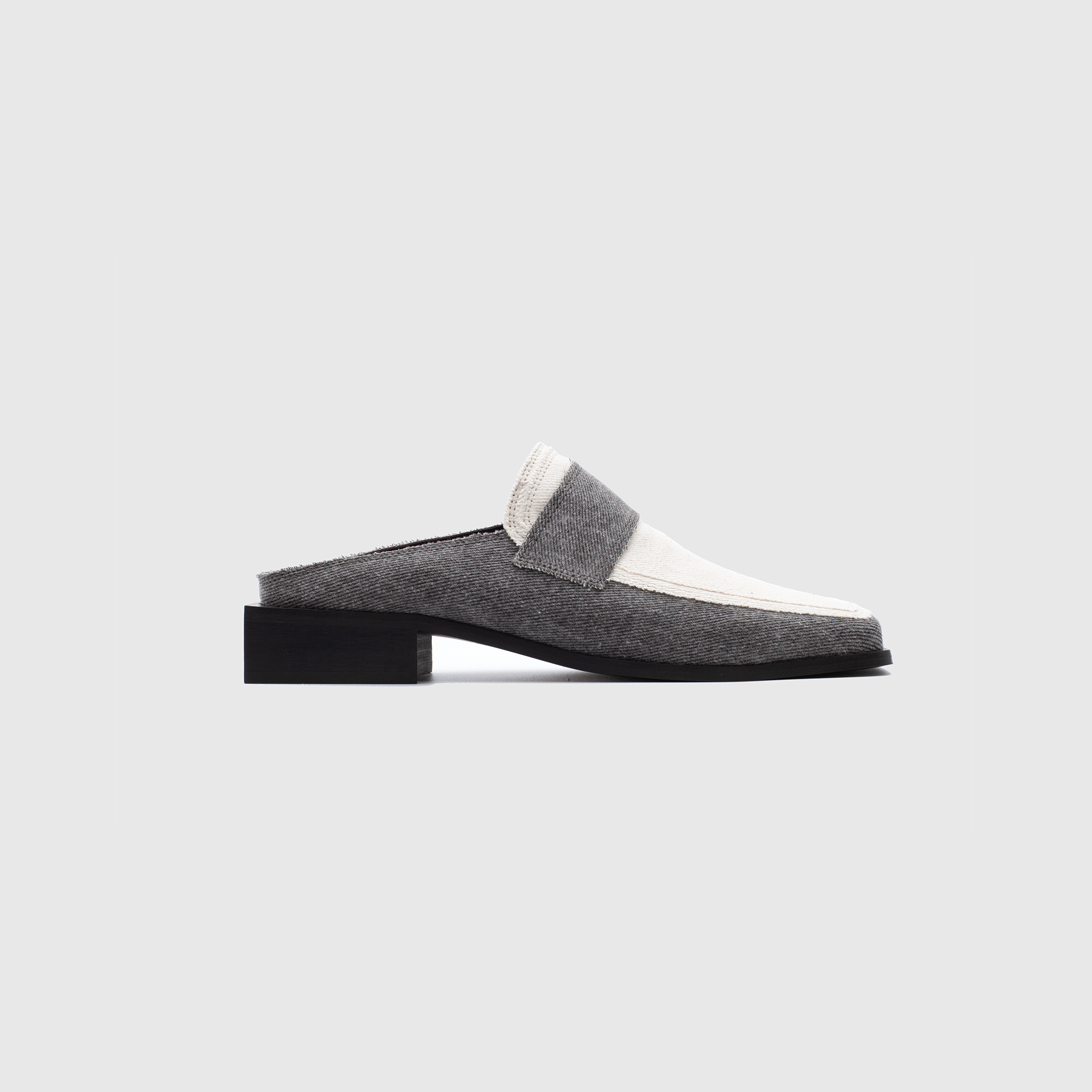 A side view of a slip-on shoe with a grey and white fabric upper reminiscent of a modern loafer. This vegan design features an open back and a low black block heel, set against a plain light grey background.