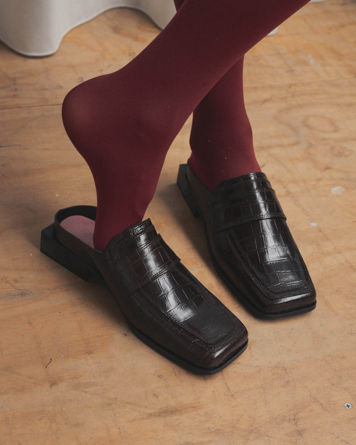 Woman in burgundy red tights wearing dark brown croco square toe loafers with square heel on a light wooden floor, left shoe slipped off showing the pink insole.